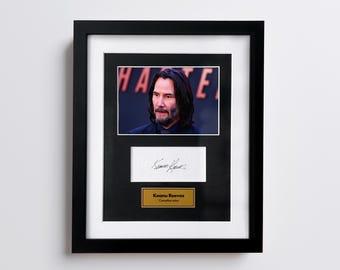 May include: Framed black and white photo of Keanu Reeves with a signature and a gold nameplate. The photo is in a black frame with a white mat.
