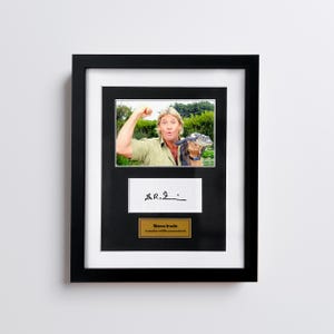 May include: Framed photograph featuring Steve Irwin holding a small crocodile. The frame is black with a white mat. Below the photo is a white rectangle with a signature and a gold nameplate that reads "Steve Irwin Australian wildlife conservationist."