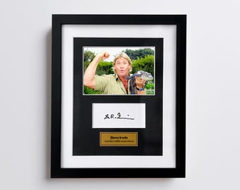 May include: Framed photograph featuring Steve Irwin holding a small crocodile. The frame is black with a white mat. Below the photo is a white rectangle with a signature and a gold nameplate that reads "Steve Irwin Australian wildlife conservationist."