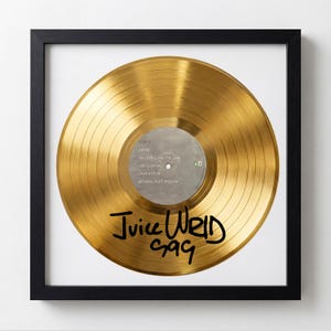 Peut inclure: Disque d'or encadré dans un cadre noir. Le disque présente un centre argenté avec des titres de chansons et des informations sur l'artiste. La signature de l'artiste, "Juice WRLD 999", est écrite au marqueur noir sur le disque.