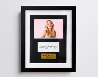 May include: Framed photograph of Sydney Sweeney with a black frame, white matting, and a gold nameplate. The photograph is of a woman with blonde hair wearing a blue and white top. Below the photo is a white card with a signature. The nameplate reads "Sydney Sweeney, American actress."