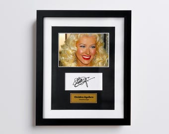 May include: Framed photograph of Christina Aguilera with a signature and a plaque that reads "Christina Aguilera, American singer". The photo shows a smiling woman with blonde curly hair and red lipstick. The frame is black.
