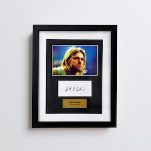 Puede incluir: Retrato enmarcado de Kurt Cobain con su firma y una placa dorada que dice "Kurt Cobain, American musician". El retrato está en un marco negro con un paspartú blanco.