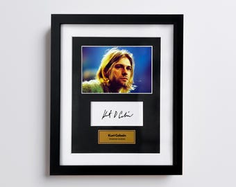 May include: Framed portrait of Kurt Cobain with his signature and a gold plaque that reads "Kurt Cobain, American musician". The portrait is in a black frame with a white mat.