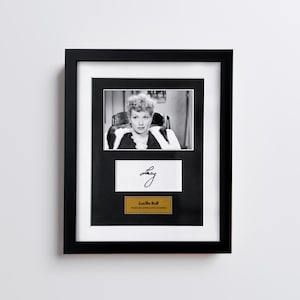 Pode incluir: Fotografia a preto e branco emoldurada em preto de Lucille Ball, com a sua assinatura e uma placa de identificação. A fotografia é montada sobre um fundo branco. A placa de identificação diz "Lucille Ball, atriz e comediante americana."
