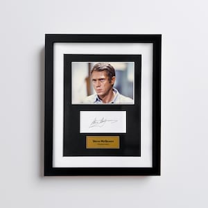 Può includere: Fotografia incorniciata di Steve McQueen, con cornice nera e passe-partout bianco. Sotto, una carta bianca con una firma e una targa dorata con la scritta "Steve McQueen, attore americano."
