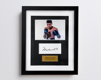 May include: Framed photograph of Muhammad Ali in boxing gloves, with his signature and a gold plaque. The black frame surrounds a white matting, showcasing the image of the boxer.