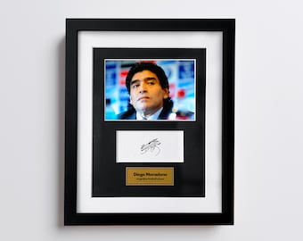 May include: Framed portrait of Diego Maradona, with a signature and a gold plaque. The black frame surrounds a photo of the Argentinian football player. Below the photo is a white rectangle with a signature, and a gold plaque that reads "Diego Maradona, Argentine football player."