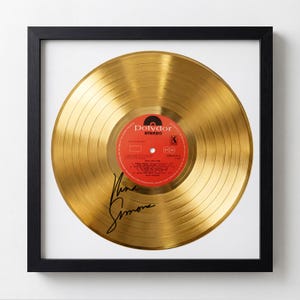 May include: A framed gold record with a red and orange label. The record has a signature in black ink and is set against a white background within a black frame. The record is a gold-colored vinyl.