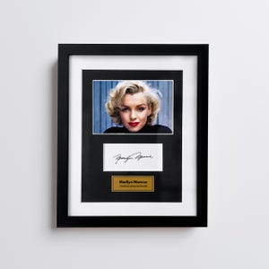 Może przedstawiać: Czarna ramka zawierająca portret Marilyn Monroe z blond włosami i czerwoną szminką, białą kartkę z podpisem czarnym tuszem oraz złotą tabliczkę z napisem Marilyn Monroe American actress and model.