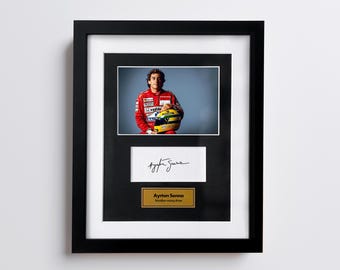 May include: Framed photograph of Ayrton Senna in a red racing suit, holding a helmet. Below the photo is a white rectangle with a signature and a gold plaque that reads "Ayrton Senna, Brazilian racing driver."