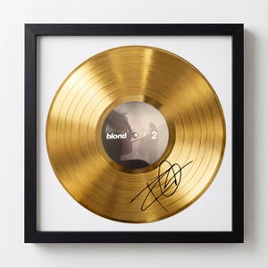May include: Framed gold-coloured vinyl record. The record features the text "blond" and "2" on the label. A black signature is on the record. The record is displayed in a black frame with a white background.
