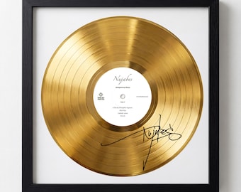May include: Framed gold-colored vinyl record with the artist's name "Nujabes" and signature. The record is displayed on a white background within a black frame. The record has a shiny, reflective surface.