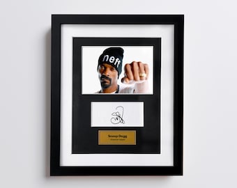 May include: Framed photo of Snoop Dogg wearing a black beanie with "neff" printed on it, clenching his fist. Below the photo is a white card with a signature and a gold plaque that reads "Snoop Dogg, American rapper."