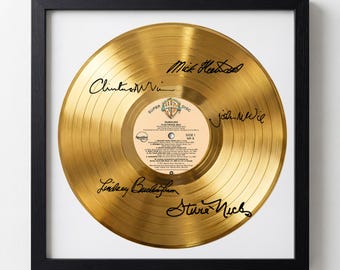 May include: A framed gold record of Fleetwood Mac's album, Rumours, with signatures. The record is gold-colored with a white and tan label in the center. The frame is black and square, with a white background.