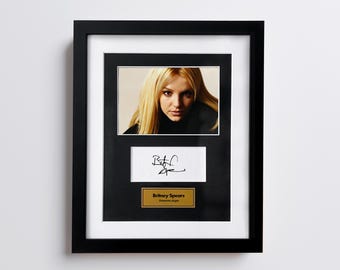 May include: Framed photograph of Britney Spears with a signature and a gold plaque that reads "Britney Spears, American singer". The frame is black with a white matting.