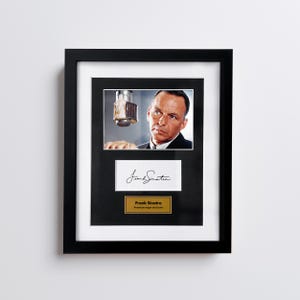 May include: Framed black and white photograph of Frank Sinatra with a microphone. Below the photo is a white card with his signature and a gold plaque that reads "Frank Sinatra, American singer and actor."