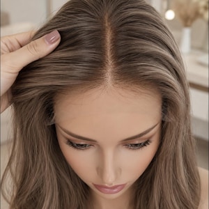 Op de afbeelding: Een lichtbruine pruik met een middenscheiding en een zichtbare lace front haarlijn. Het haar heeft subtiele highlights en een golvende textuur. De tekst "our wigs hairline" staat in het wit onderaan in het midden van de afbeelding.