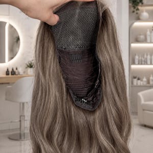 Op de afbeelding: Een lange, golvende, asbruine pruik met een donkere kanten kapconstructie, interne kammen en verstelbare bandjes. De onderkant van de kap is zichtbaar, met het gaasmateriaal en de bevestigingspunten. Tekst 'our wigs cap' onderaan.