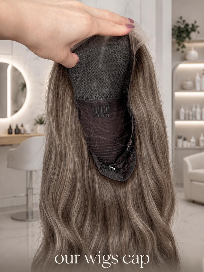 Op de afbeelding: Een lange, golvende, asbruine pruik met een donkere kanten kapconstructie, interne kammen en verstelbare bandjes. De onderkant van de kap is zichtbaar, met het gaasmateriaal en de bevestigingspunten. Tekst 'our wigs cap' onderaan.