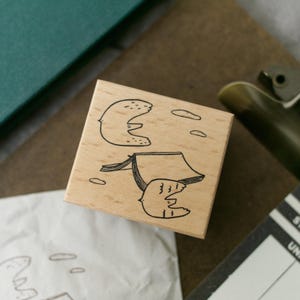 May include: A wooden stamp with a whimsical design featuring two bird-like figures and several small, oval shapes. The stamp's design is in black ink on a light-coloured wood block. The stamp is square and has a simple, clean design.