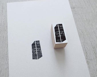 May include: A wooden stamp with a black and white design, next to a stamped impression of the same design. The stamp has a rectangular shape with a detailed graphic and text. The stamped image is a stylized geometric shape.
