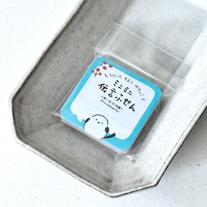 May include: A pack of square sticky notes with a light blue border and a cartoon bird illustration. The notes are in a clear plastic sleeve and are on a grey ceramic tray. Japanese text is visible on the notes.