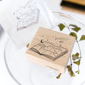 May include: A wooden stamp featuring a black ink illustration of an open book with mountains, stars, a crescent moon, and a planet. A stamped image on a white surface is also visible.