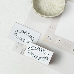 Op de afbeelding: Twee witte visitekaartjes met een zwarte ovale stempel met de tekst "E. Garguel". Een kleine, lichtgekleurde kom en een papier met een huisontwerp zijn ook zichtbaar. De kaarten hebben een vintage esthetiek, met focus op design.