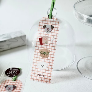 May include: A clear bookmark with a grid pattern and illustrations of dogs wearing glasses, a book, and a pen. The word "DOGS" is printed at the bottom. A green ribbon is attached at the top.