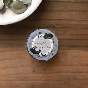 May include: A roll of washi tape with a light blue background. The tape features black illustrations of cats, flowers, and musical notes. The text "Twilight cat and flowers" is written on the tape in cursive.