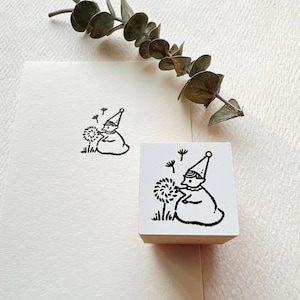 May include: A square wooden rubber stamp featuring a black ink illustration of a small figure in a pointed hat blowing on a dandelion, with a matching stamped image on the paper below and a sprig of dried eucalyptus leaves positioned above.