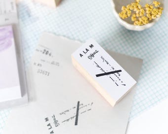 May include: A white rectangular stamp with the text "A LA M" and other script, crossed out with a black line. The stamp is on a piece of paper with a similar stamped design. Other stationery items are in the background.