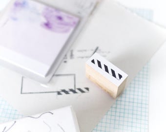 May include: A wooden stamp with a white top featuring a black diagonal stripe design. The stamp is next to a stack of white paper, a pad of purple floral sticky notes, and a small package with a line drawing.