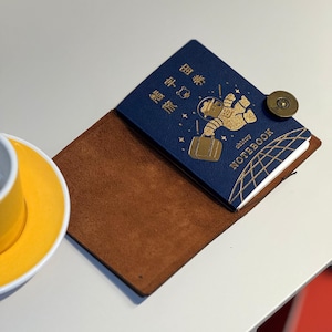 Peut inclure: Un carnet bleu foncé avec un motif d'astronaute en feuille d'or, le texte "obltoy NOTEBOOK" et des caractères chinois. Le carnet est inséré dans une couverture en cuir marron fixée par un bouton-pression métallique couleur bronze.