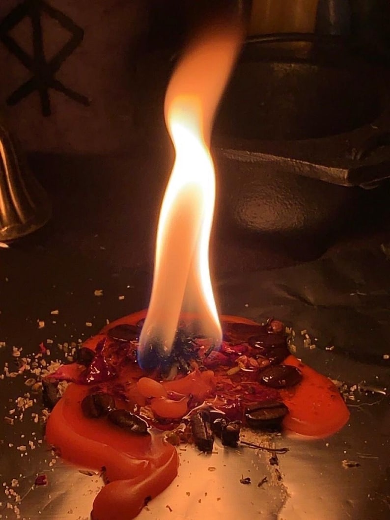 May include: A lit candle with a bright orange flame. The candle is surrounded by red wax, herbs, and other small items. The background is dark, with a symbol visible on the left side.