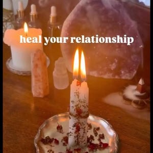 May include: Two white taper candles bound together with twine, decorated with dried flower petals, and placed on a silver-toned scalloped plate. The candles are lit, and the text "heal your relationship" is centered in the image.
