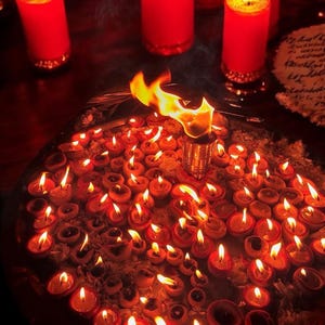 May include: A collection of small, lit oil lamps and several large red candles with golden bases. The lamps are arranged on a round tray, creating a warm, inviting glow. A flame burns brightly from a central metal holder.
