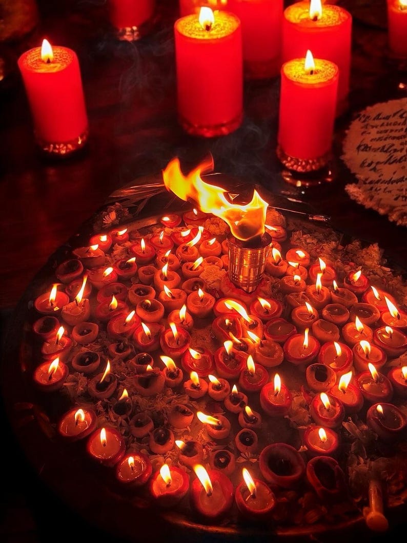 May include: A collection of small, lit oil lamps and several large red candles with golden bases. The lamps are arranged on a round tray, creating a warm, inviting glow. A flame burns brightly from a central metal holder.