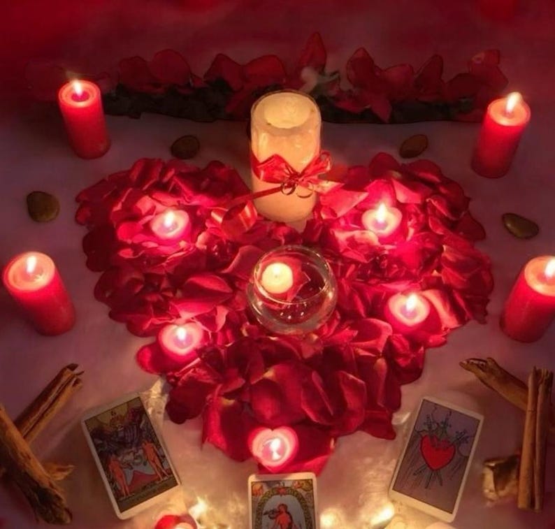 May include: A romantic scene with lit candles and red rose petals arranged in a heart shape. A large pillar candle is tied with a red ribbon. Tarot cards and cinnamon sticks are also present, creating a mystical ambiance.