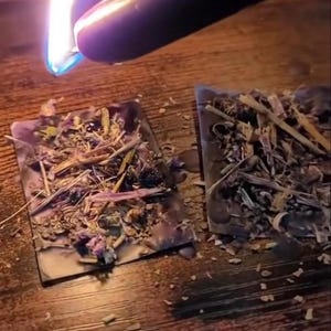 May include: A lit black candle flame above two small, square, dried herb bundles. The bundles are filled with various dried herbs and flowers. The background is a dark wooden surface.
