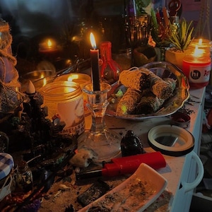 May include: A dimly lit scene featuring lit candles, jars, and a silver platter with wrapped bundles. A black candle burns in a glass holder, and a red lighter and ashtray are visible. The setting suggests a spiritual or ritualistic theme.