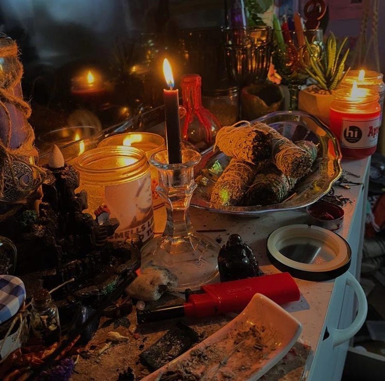 May include: A dimly lit scene featuring lit candles, jars, and a silver platter with wrapped bundles. A black candle burns in a glass holder, and a red lighter and ashtray are visible. The setting suggests a spiritual or ritualistic theme.
