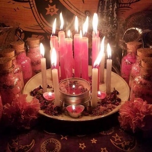 May include: A spiritual display with lit pink and white candles on a plate, surrounded by small jars with cork stoppers, and pink flowers. A skull and tapestry are in the background, creating a mystical atmosphere.