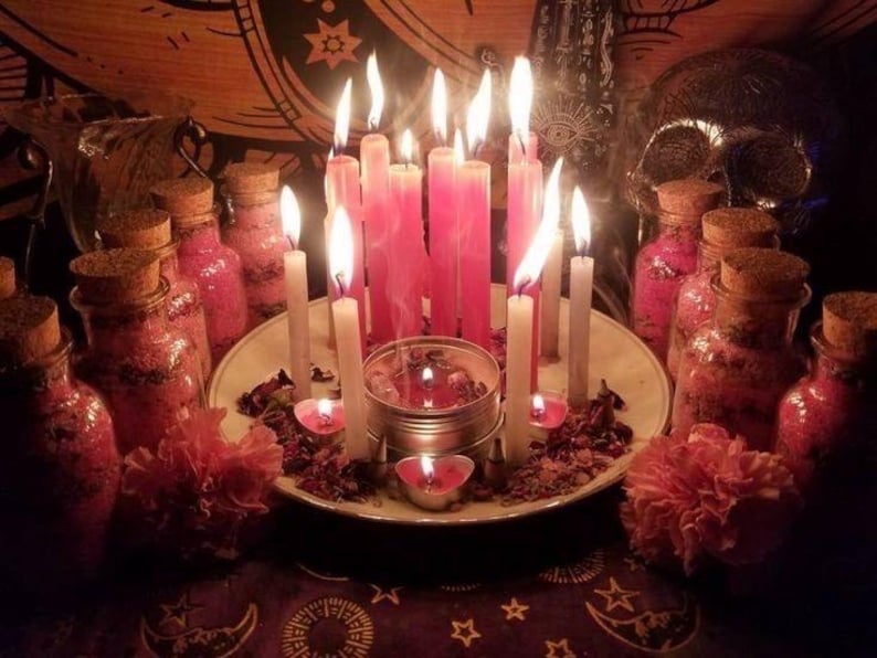 May include: A spiritual display with lit pink and white candles on a plate, surrounded by small jars with cork stoppers, and pink flowers. A skull and tapestry are in the background, creating a mystical atmosphere.