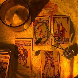 May include: A candle in a glass jar and several tarot cards are arranged on a textured surface. The cards feature illustrations and text in a foreign language. Incense sticks and a decorative metal incense holder are also present.