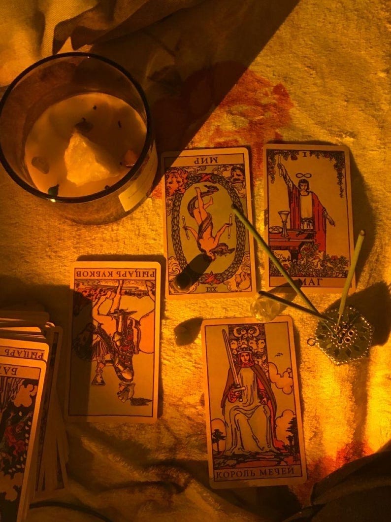 May include: A candle in a glass jar and several tarot cards are arranged on a textured surface. The cards feature illustrations and text in a foreign language. Incense sticks and a decorative metal incense holder are also present.