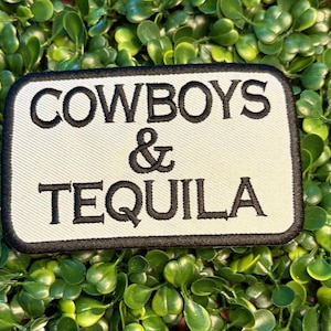 May include: A rectangular patch with the words "COWBOYS & TEQUILA" embroidered in black on an off-white background. The patch has a black border and is set against a backdrop of green foliage.