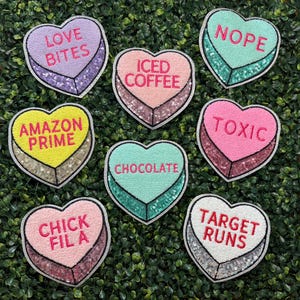 May include: A collection of heart-shaped patches with sequin borders and various phrases. The patches feature phrases like "LOVE BITES," "ICED COFFEE," "NOPE," "AMAZON PRIME," "CHOCOLATE," "TOXIC," "CHICK FIL A," and "TARGET RUNS."