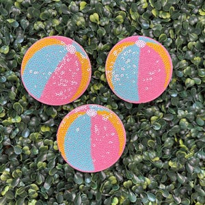 May include: Three round, sequined beach ball-themed coasters. Each coaster is divided into three sections: pink, blue, and yellow. The coasters are set against a backdrop of green foliage.